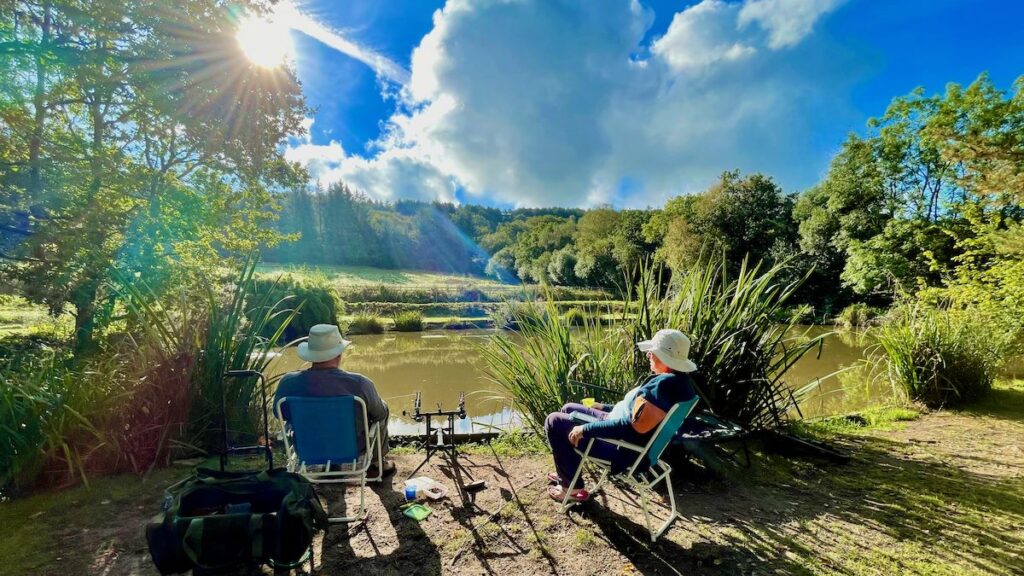Tranquil view of a fishing pond at South Farm Holiday Cottages and Fishery. Two people enjoy a sunny afternoon fishing, surrounded by lush greenery under a blue and cloudy sky in the UK.