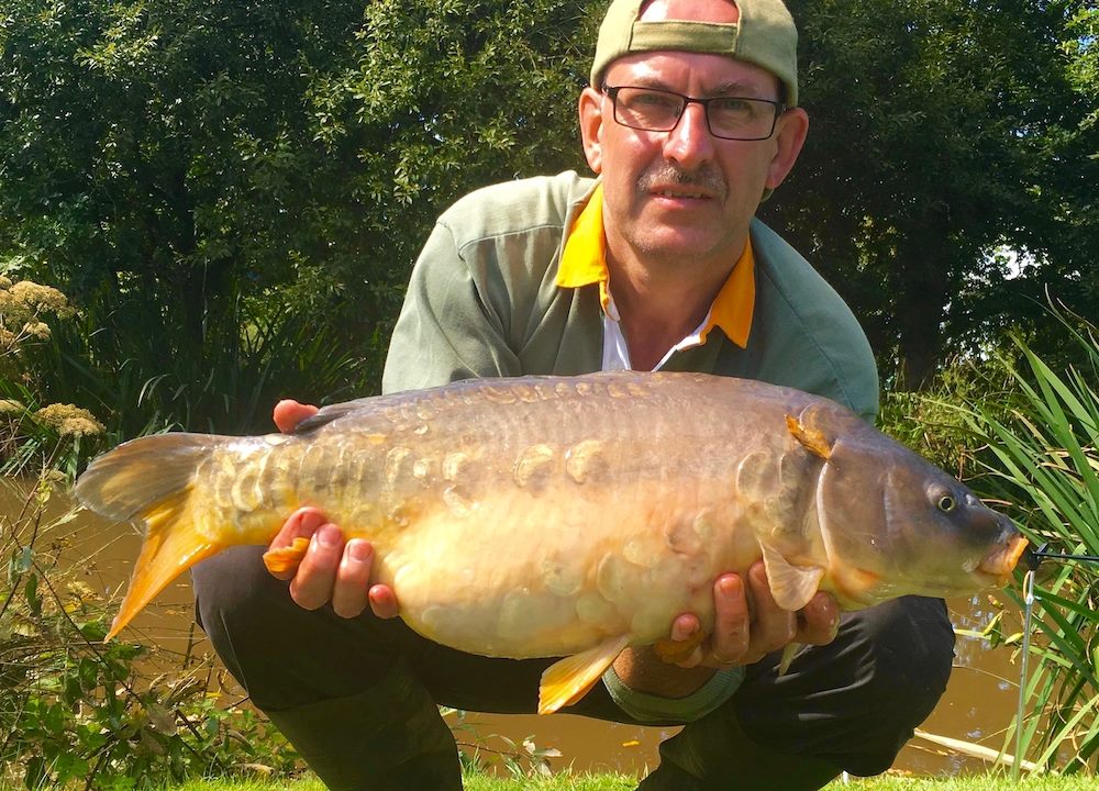 angler in glasses and a cap kneels fishing lake, holding a large common carp he has caught.