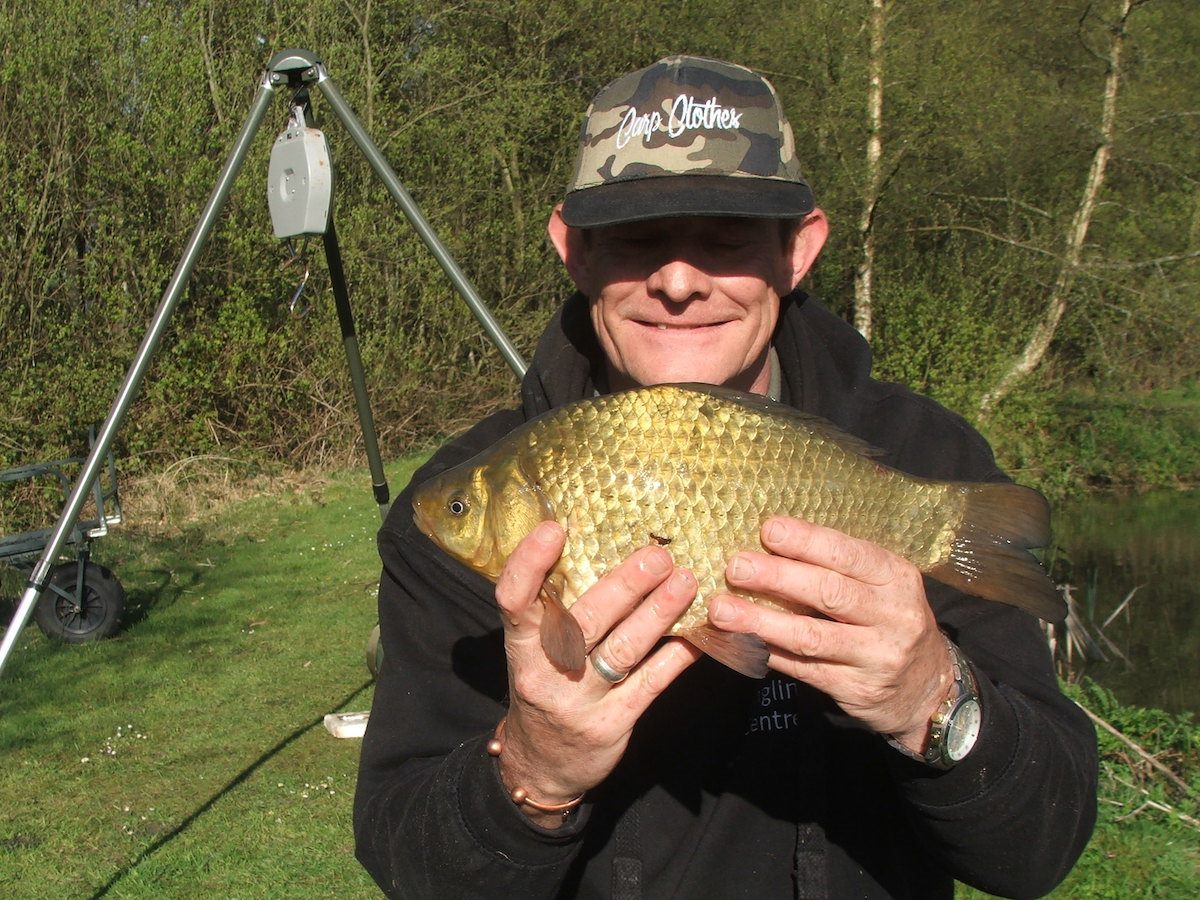 smiling angler in a camouflage cap holds a golden carp he has caught at Lower Ram Lake. He is positioned by the water's edge with fishing gear visible in the background, including a rod pod and bite alarm.