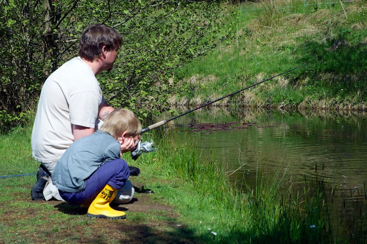 Father and son enjoying a fishing outing at Upper Ram Lake. The father is actively fishing, and his son, wearing bright yellow wellies, is engaged in the activity.