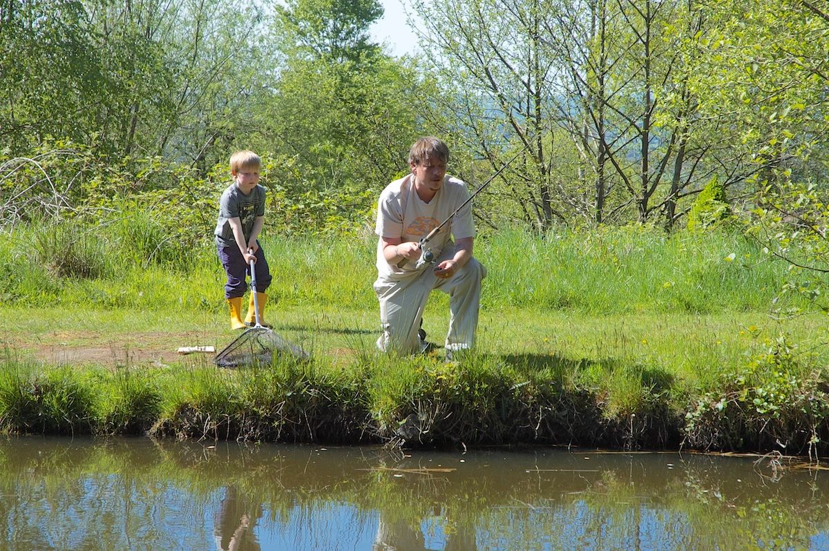 A father and son enjoying a fishing trip at Upper Ram lake. The father is actively fishing while his son observes with a net, amidst the lush green vegetation and trees.