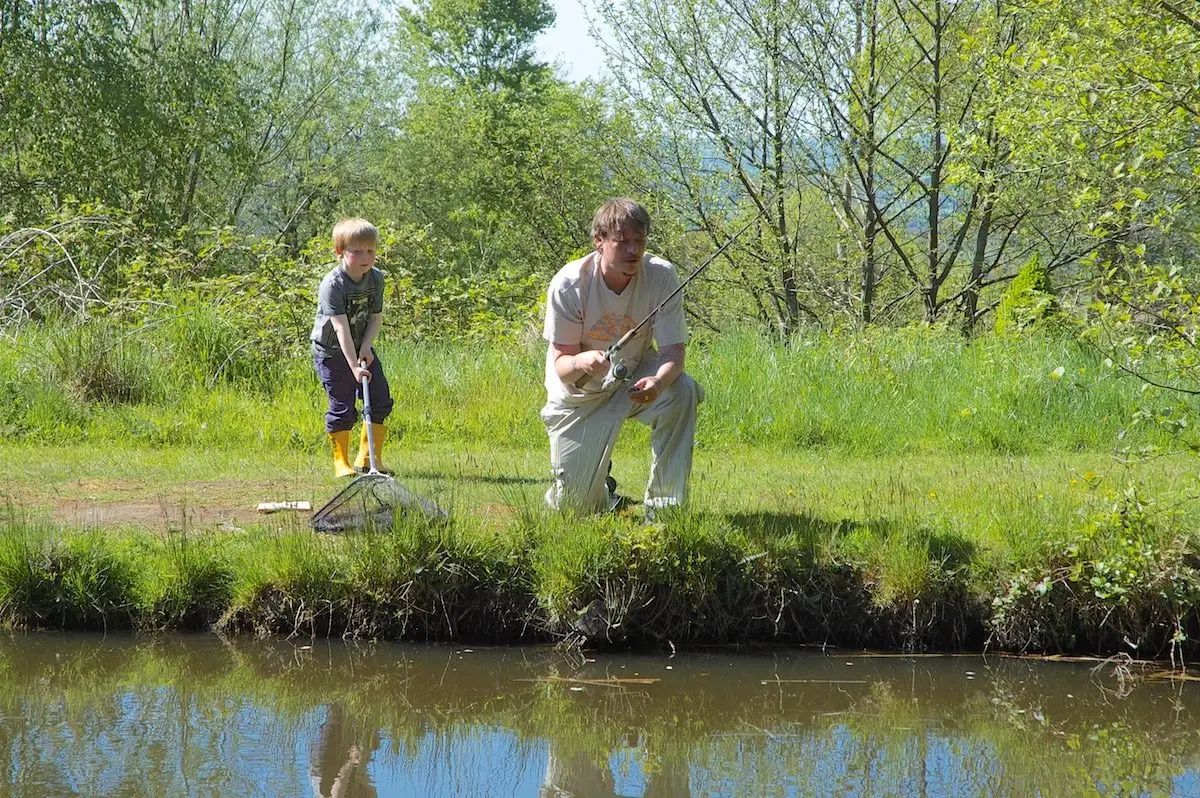 A father and son enjoying a fishing trip at Upper Ram lake. The father is actively fishing while his son observes with a net, amidst the lush green vegetation and trees.